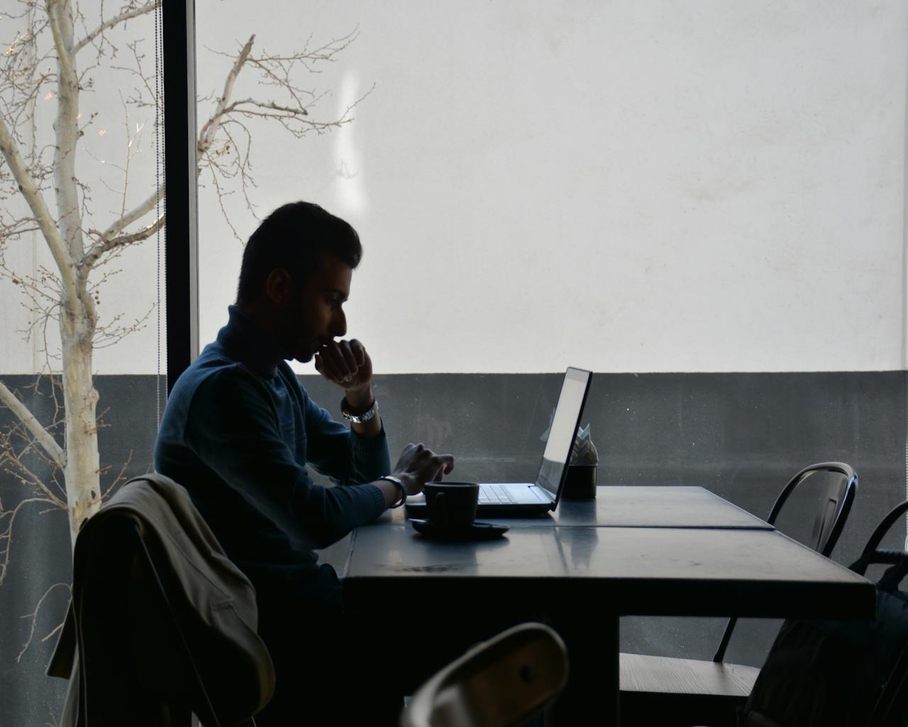 Silhouette of a man concentrating on his laptop while sitting at a café table.