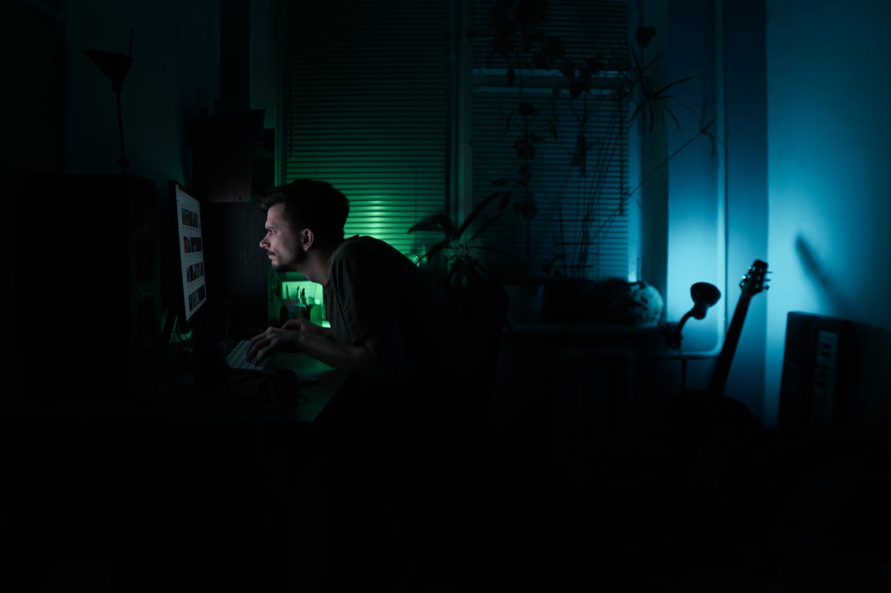 A man focused on work at his computer, late at night in a dimly lit home office.