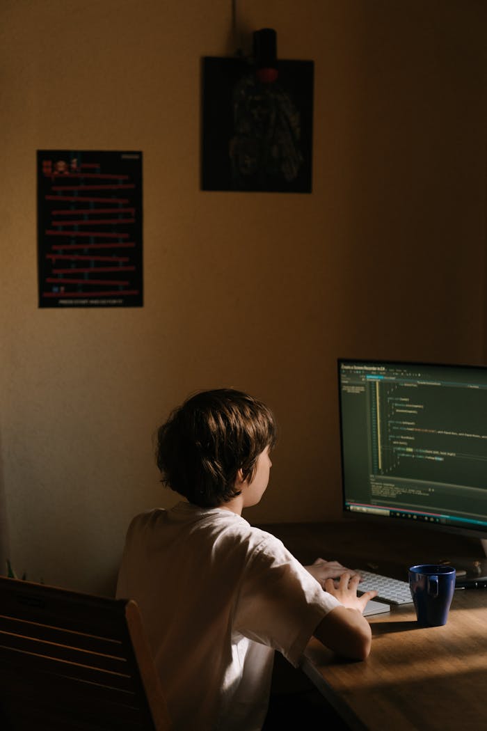 A young boy focused on coding on a computer in a dimly lit room, embodying technology and concentration.
