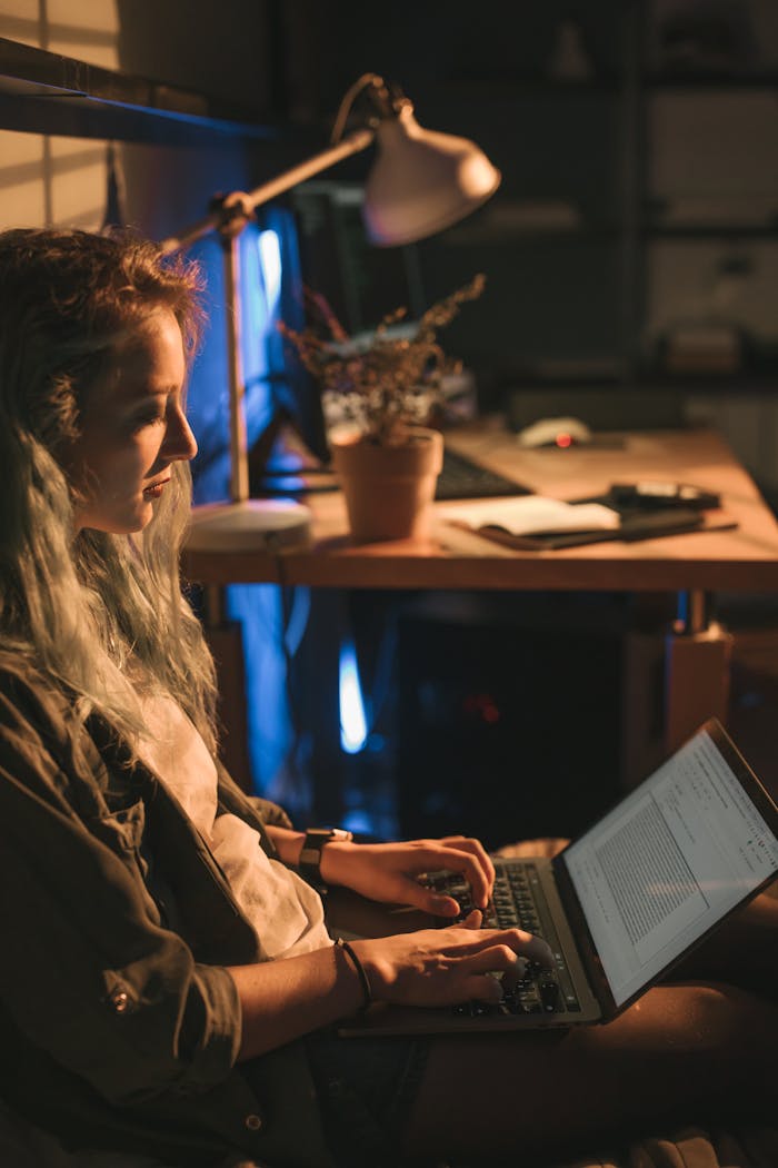Woman focused on laptop work in a cozy, dimly lit room setting, showcasing a modern home office vibe.
